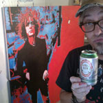 Photo of artist Mark Goodman making duck lips, holding a beer in foreground with painting of Syd Barrett in background
